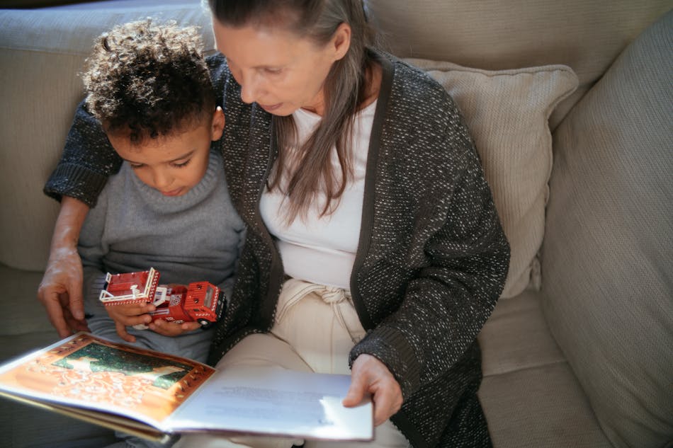 An adult and a child dressed in cosy knitted jumpers, sitting on a sofa. The adult has one arm around the child and a colourful book open in their lap. The child is holding a toy fire engine and looking down at the pages.