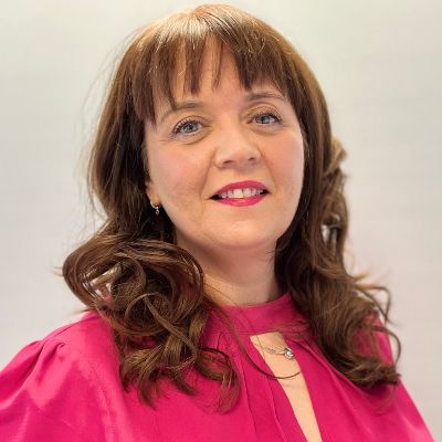 Jaqueline (CEO) smiles warmly for the camera. They are a white person with shoulder-length brown hair, wearing a pink blouse.