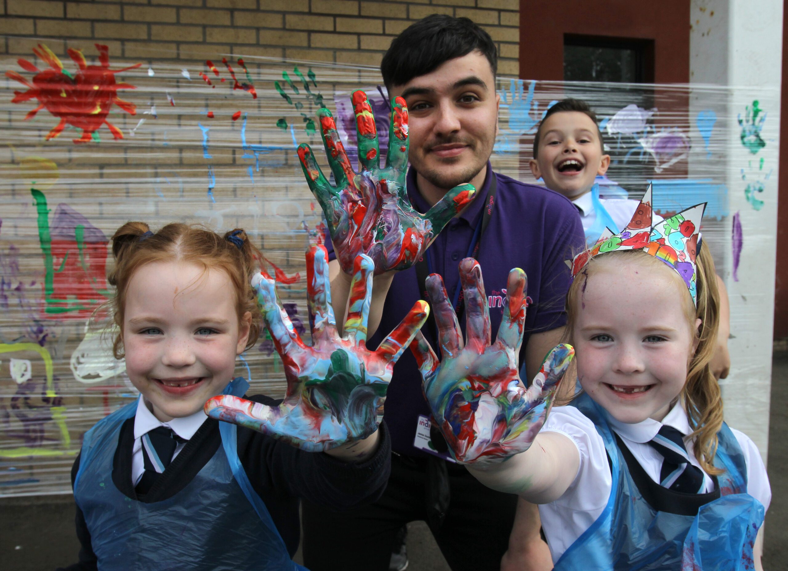 An adult and three children, happily holding up their paint-covered hands for the camera. In the background, their colourful painted artwork is displayed against a brick wall.