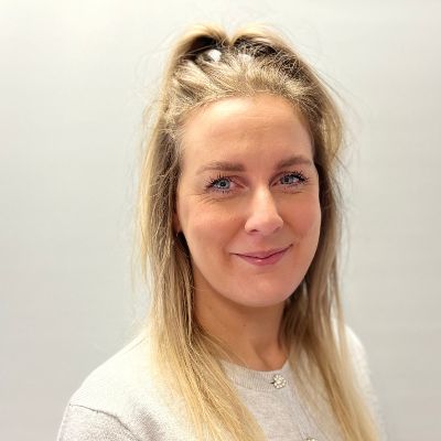 Steph smiles warmly for the camera. They are a white person with blonde hair, wearing a white long-sleeved top.