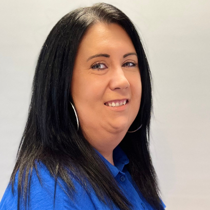 Debby Larkin smiling warmly for the camera. They are a white person with long black hair, wearing a blue polo shirt and silver hoop earrings.