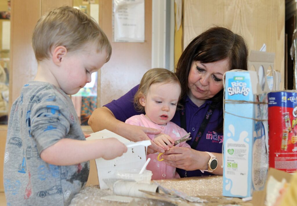 An Indigo staff member is at a table with two babies, one wearing pink clothes and the other in grey. The staff member is holding the baby in a pink bodysuit while they play together