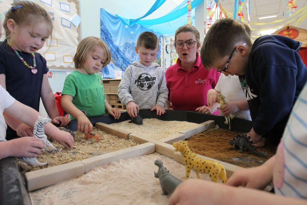 A group of children playing with toy animals at a desk that has 4 sections that have a different surface, one sawdust, sand, muesli and dry pasta