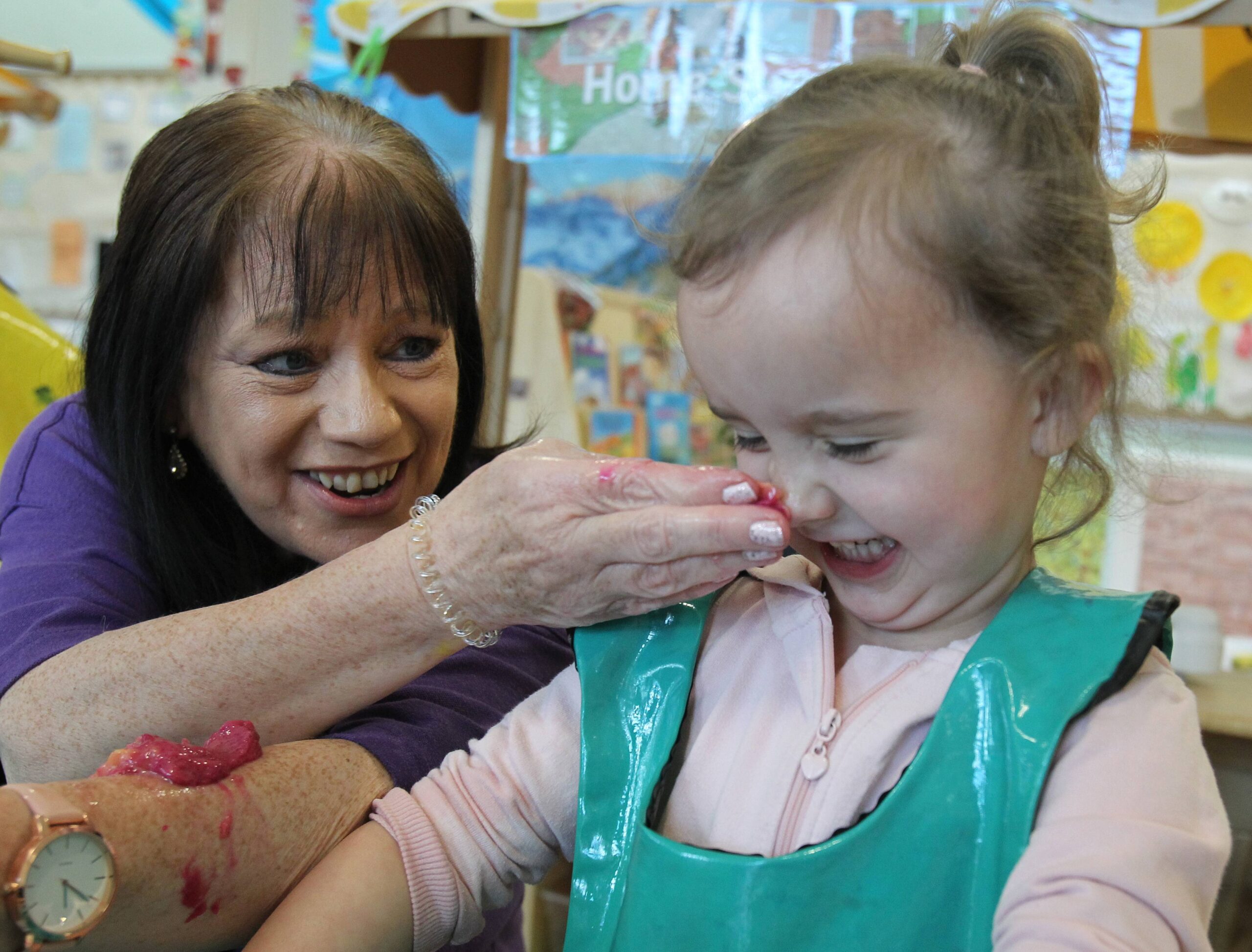 An Indigo staff member wearing a purple shirt is interacting with a child in a pink shirt and apron. They are playing with wet pink clay, gently placing it on the child’s face in a playful and teasing manner