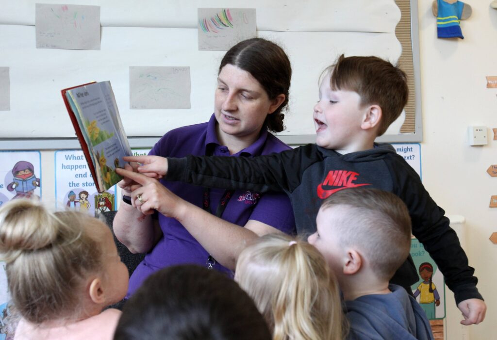 An Indigo staff member wearing a purple shirt stands holding a book while a group of children gather around, listening attentively. One child is pointing directly at the book