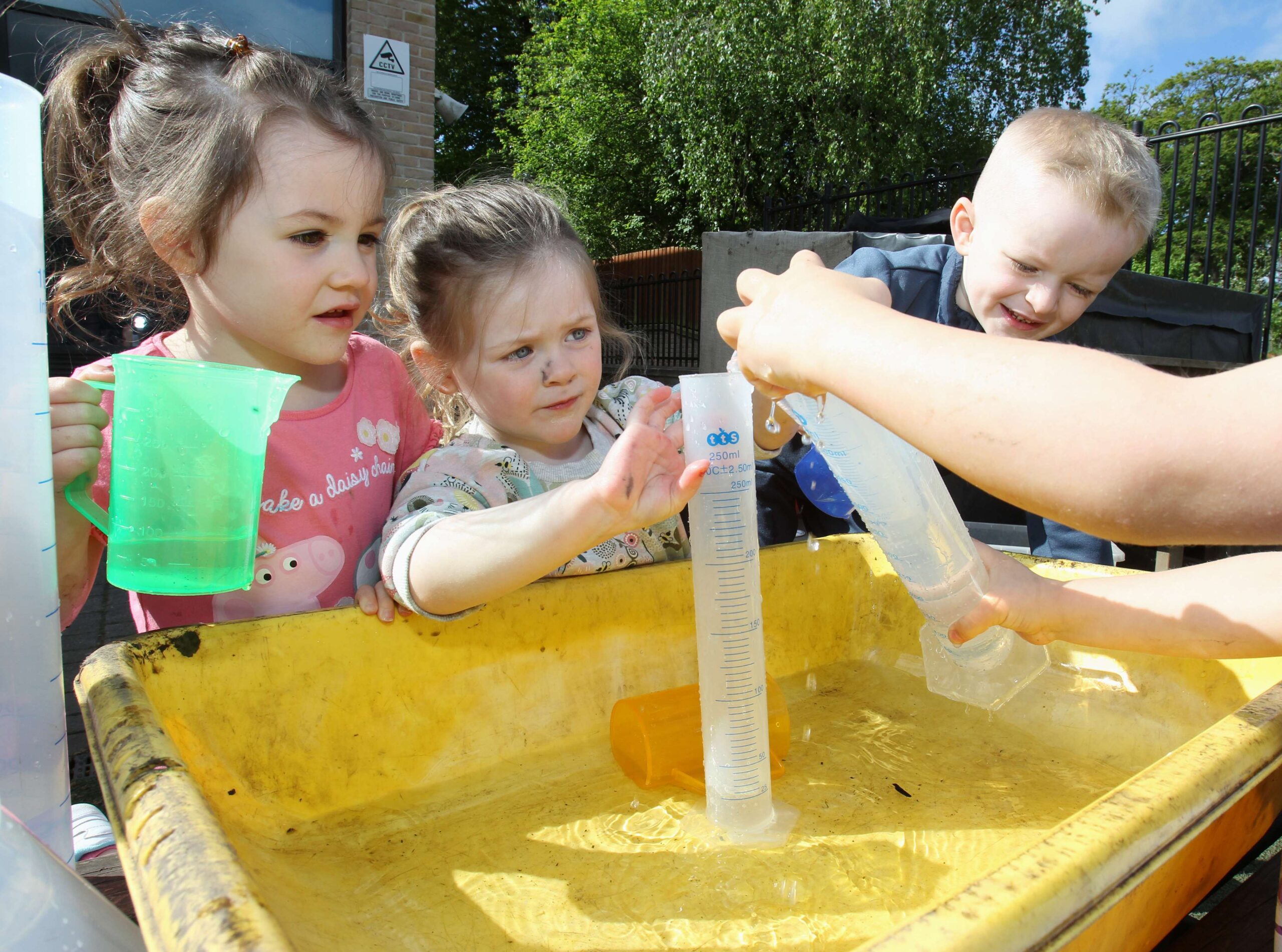 Children playing outside with a yellow tub full of water, plastic jugs and measuring cylinders