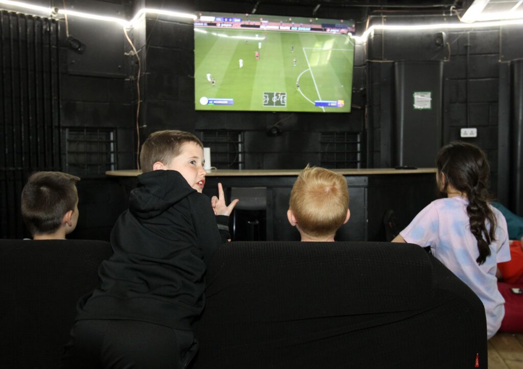 Four children are seated on black bean bags, watching football. Three of the children are dressed in black clothes, and one is wearing a purple t-shirt