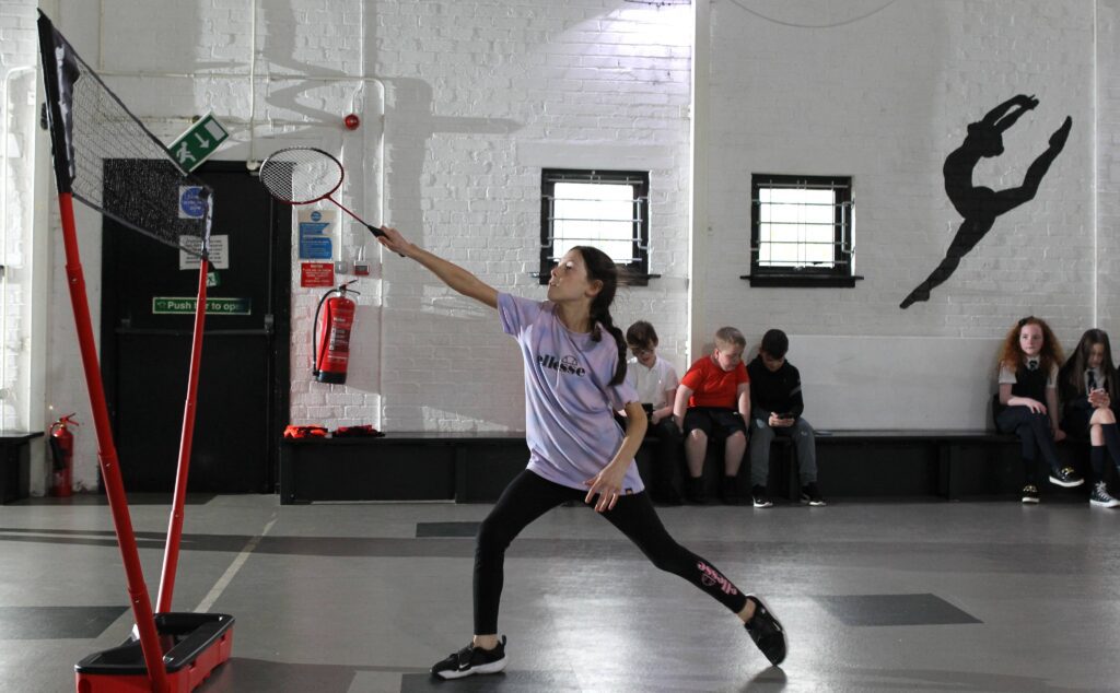 A child wearing a purple t-shirt and gym leggings is standing in a gym, holding and playing with a tennis racket