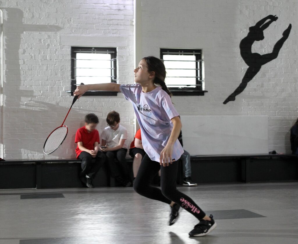 A child in a purple shirt and sports leggings holding a racket, playing in a gym