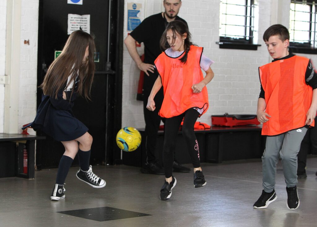 Three children in a sports gym playing football. Two are wearing orange vests and sports clothes, while the third is in a school uniform