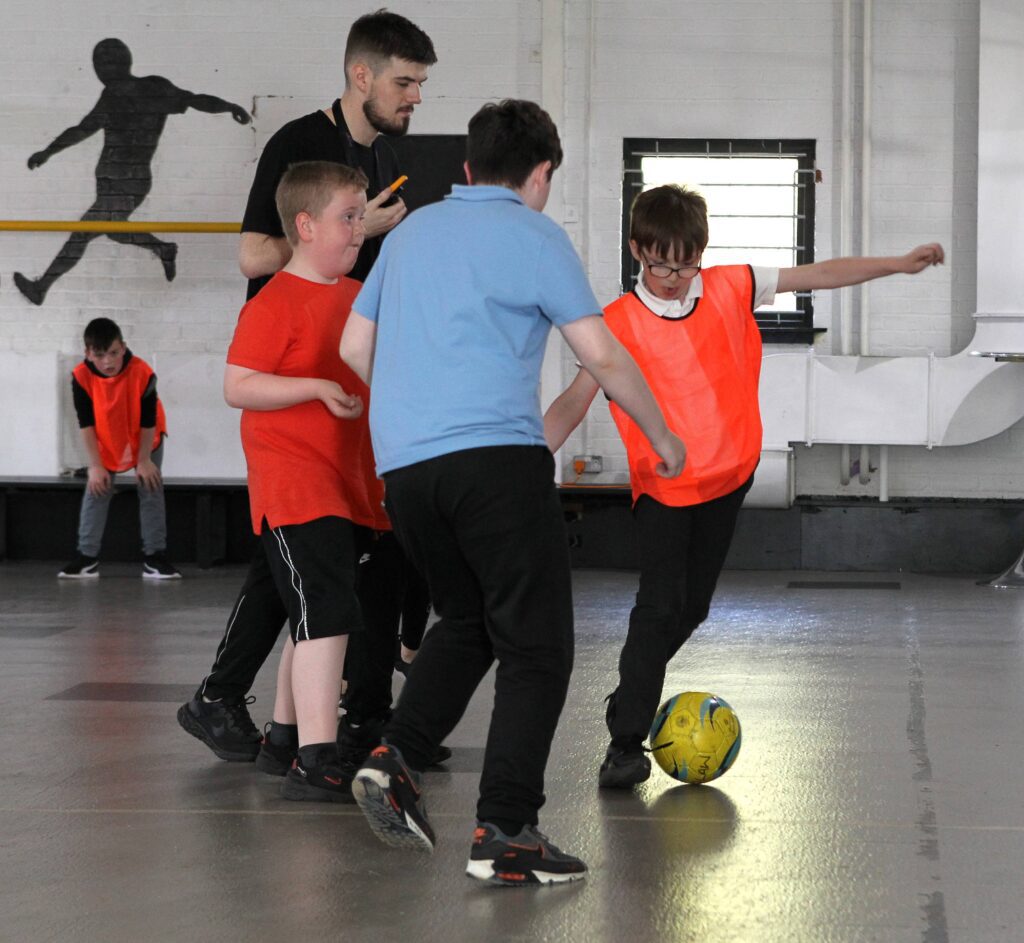 Several children are playing football in a large, bright gym with white walls