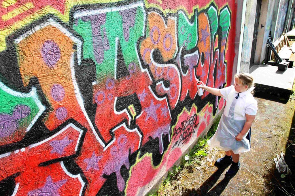 A child dressed in a school unform and wearing a protective apron and glasses, standing outdoors in the sunlight. They are using a can of spray paint to paint the wall in front of them, which is decorated with red, black, green, orange, and blue lettering.