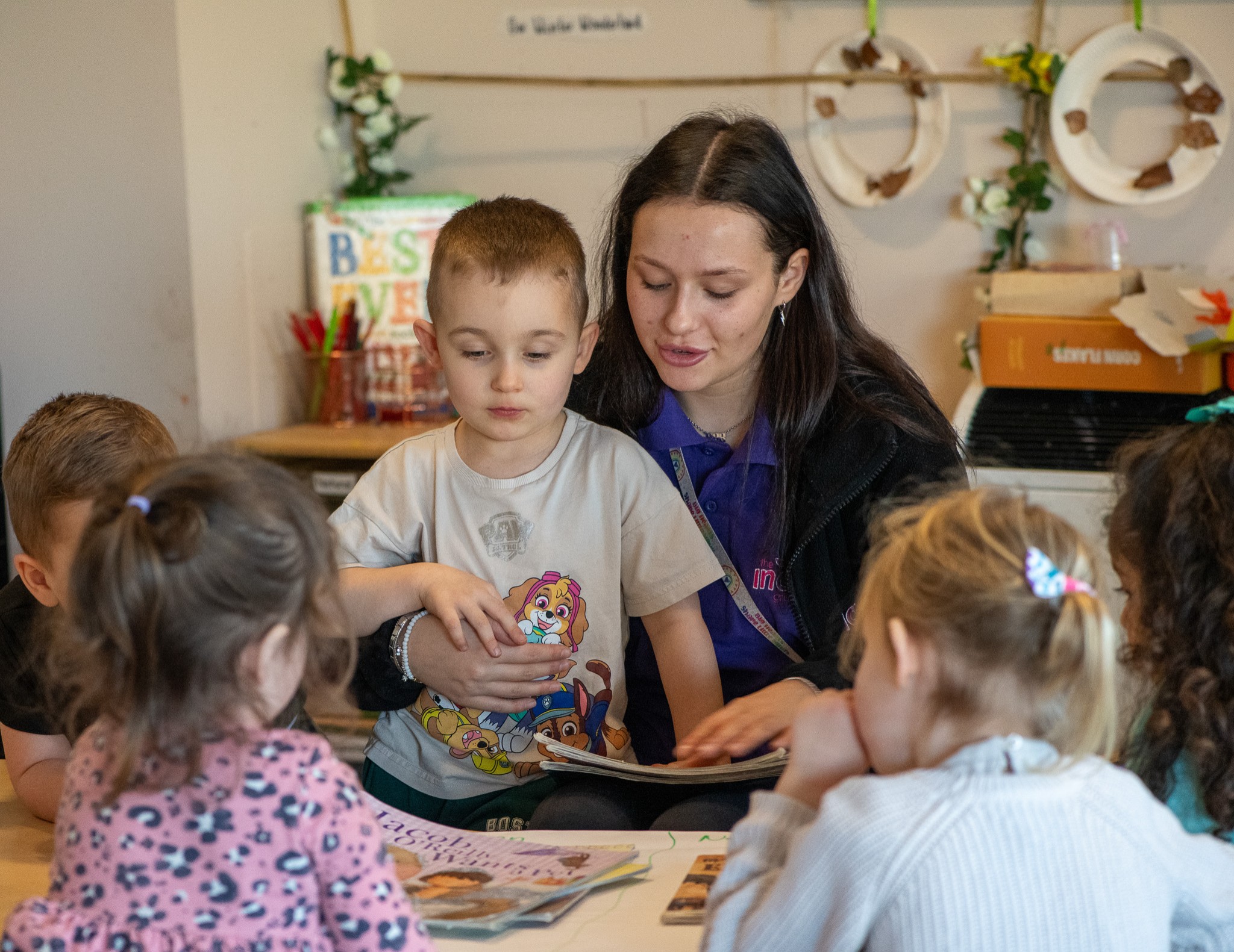 An adult dressed in a blue polo shirt, sitting with one hand on a stack of books in their lap and the other hand around a child standing close beside them. Four children are sitting in front of them, gathered around colourful picture books on a table.