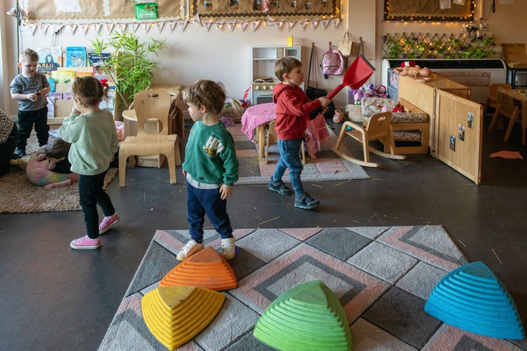 Five children using an indoor space with a rack of childrens' books, rugs, wooden chairs, and toys. One child is lying on a bean bag chair, and four children are standing, one playing with a dustpan.