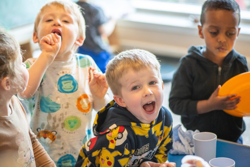Four children looking at the camera. Arts and crafts materials are on the table in front of them. The child in front is smiling widely, and the child in the back is holding a plate