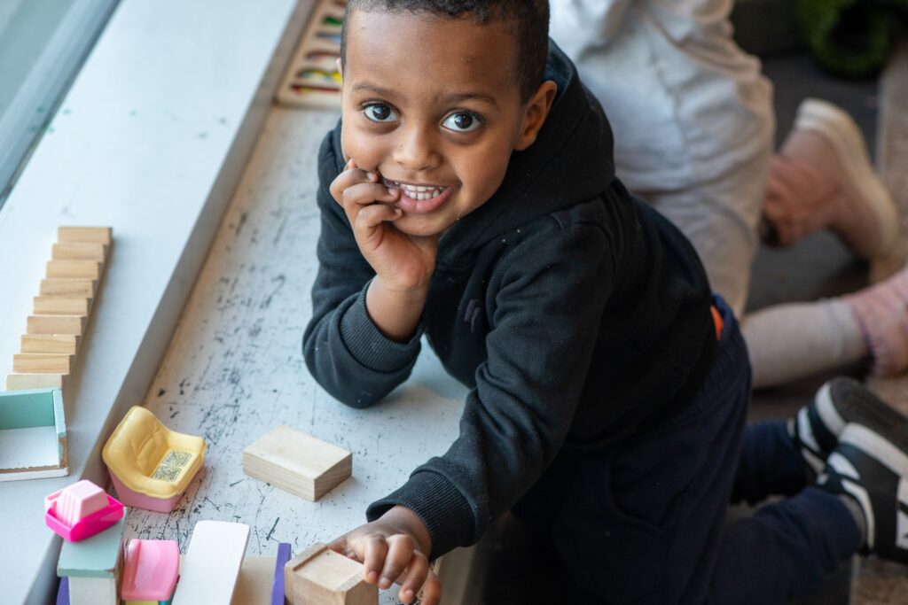 A child kneeling and smiling up at the camera. They are resting their head in one hand and playing with wooden blocks with the other.