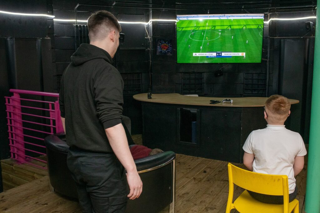 A teenager in black clothes standing and watching football, while a child in a school uniform sits in a yellow chair