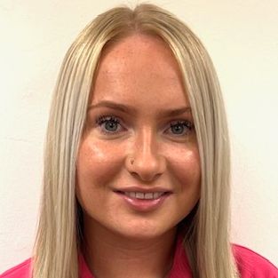 Amy Eagle smiles warmly for the camera. They are a white person with blonde hair, wearing a pink blouse