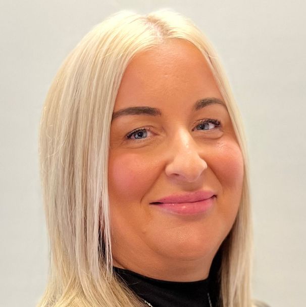 Ashley smiles warmly for the camera. They are a white person with blonde hair, wearing a black polo-neck top.