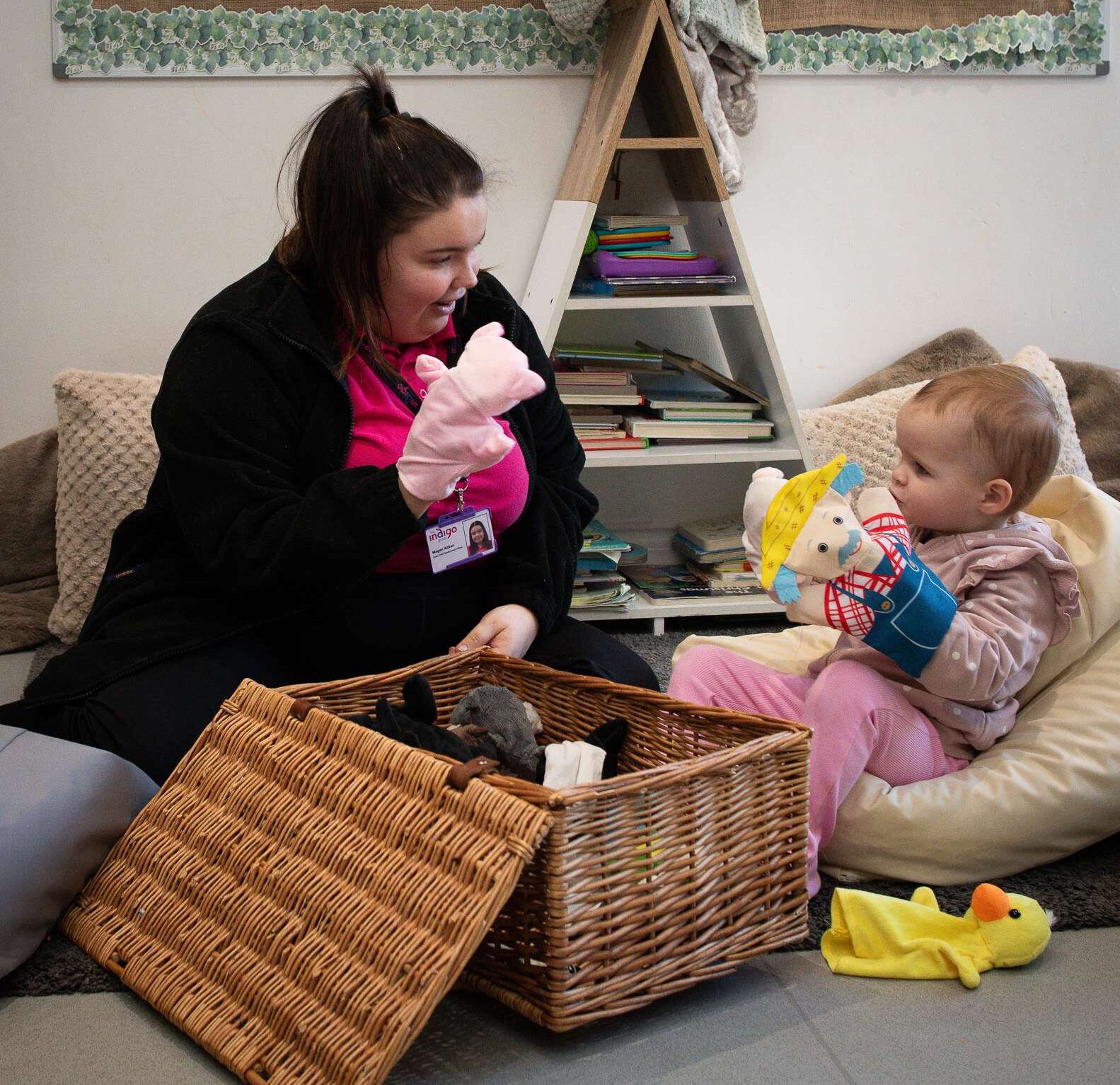 An Indigo staff member wearing a pink t-shirt and black hoodie is holding a puppet while a baby sits opposite, observing the toy with curiosity. A woven toy box is placed between them, and a triangular bookshelf with books is visible in the background
