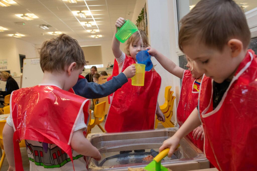 a group of young children playing with small coloured jugs of water and they are wearing a red waterproof apron. There is a small sand pit in the foreground