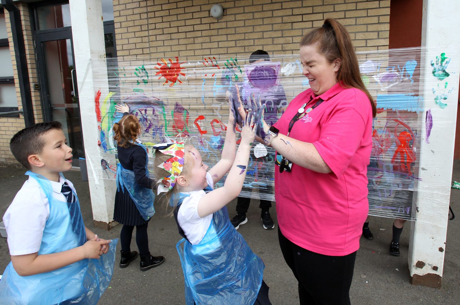 Six children and an Indigo staff member standing outdoors. The children are wearing school uniforms and plastic aprons. In the foreground, a child and the staff member give each other a high five with paint-covered hands. In the background, colourful artwork is displayed against a brick wall.
