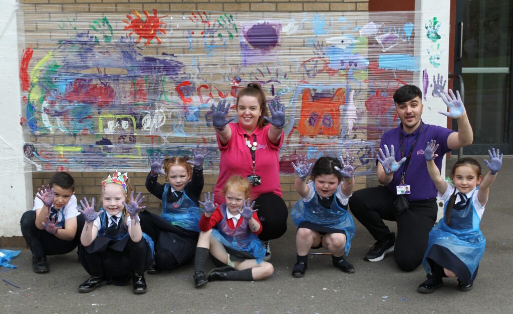 A group of six children and two adults, crouching down outdoors. The children are dressed in school uniforms and wearing plastic aprons. They are all holding up their paint-covered hands and smiling widely. In the background, their colourful painted artwork is displayed against a brick wall.
