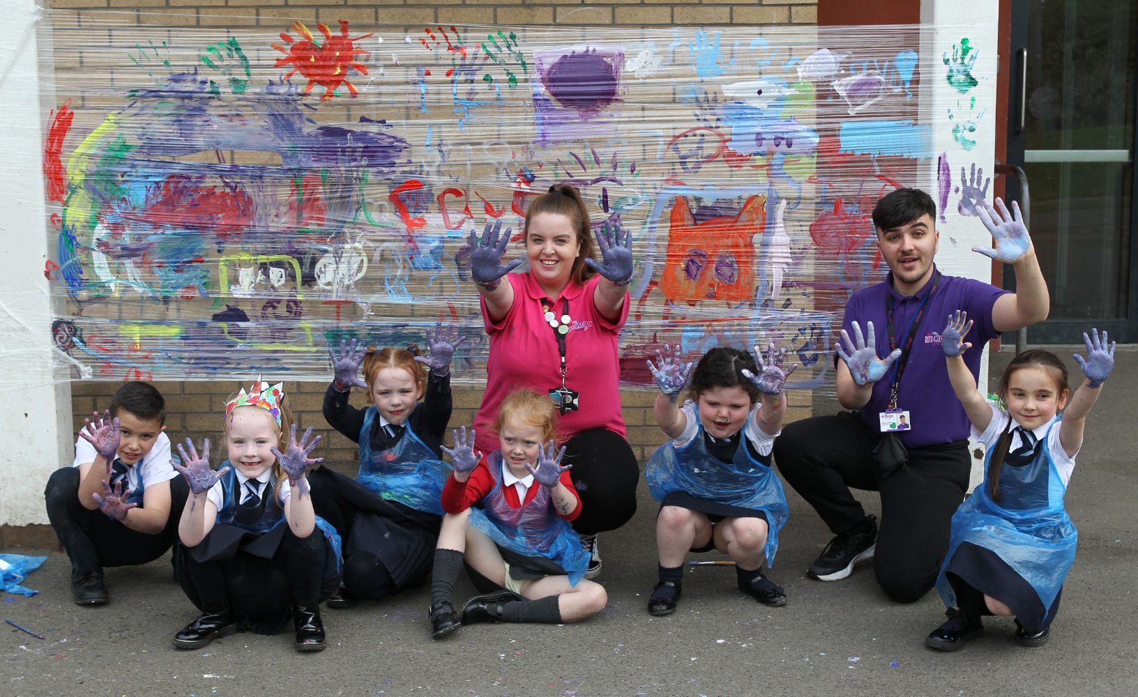 A group of six children and two adults, crouching down outdoors. The children are dressed in school uniforms and wearing plastic aprons. They are all holding up their paint-covered hands and smiling widely. In the background, their colourful painted artwork is displayed against a brick wall.