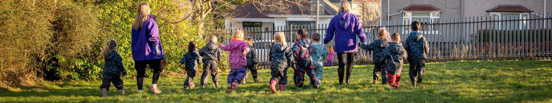 Pictured from behind, eleven children and two adults walking in a group outdoors in the sunshine with trees in the background. The adults are wearing purple jackets and the children are dressed in waterproof clothing and wellington boots.