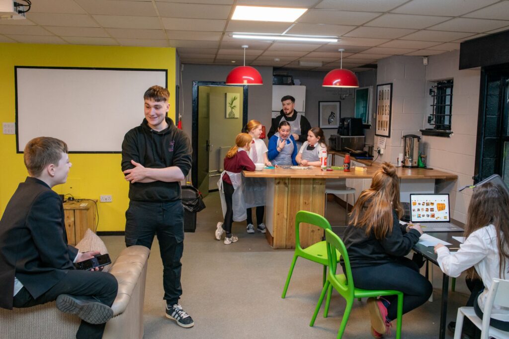 A view of a room where two teenagers in black clothing are chatting in the foreground. In the background, children wearing school aprons are preparing food, and a child sits at a table speaking with an Indigo staff member.