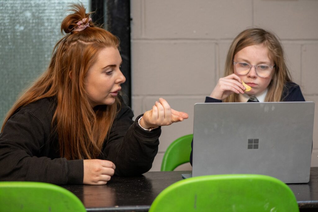 An Indigo staff member and a child in a school uniform sitting close at a table. The staff member is explaining something while pointing at a laptop, and the child is looking at the laptop screen