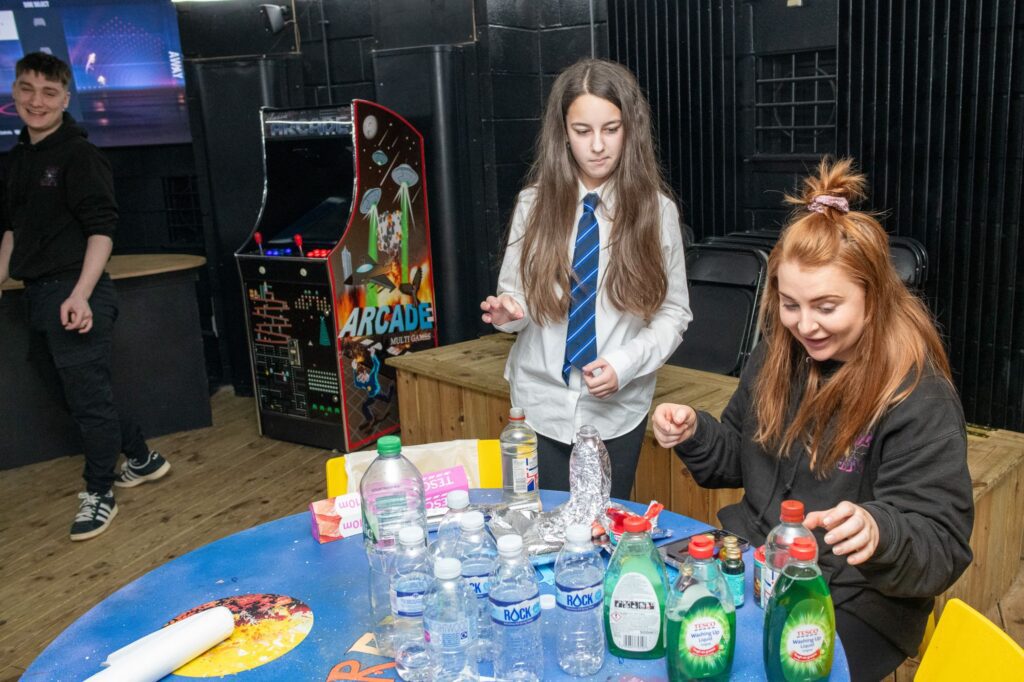 In a game room, two children—one in sports clothes and one in a school uniform—are at a round table with an Indigo staff member in black clothes, doing a science experiment. An arcade game machine is visible in the background