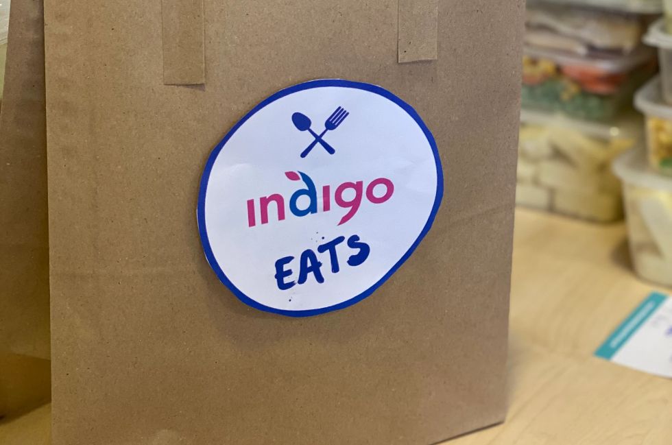 A brown paper bag with a large, white paper sticker displaying the Indigo Eats logo