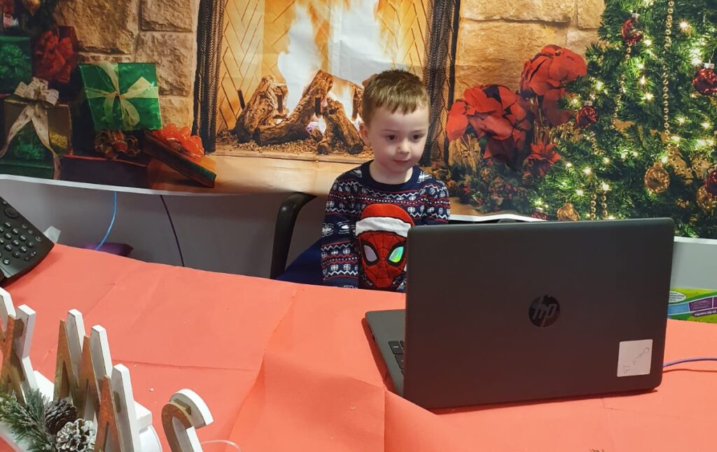 A child wearing a Spiderman Christmas jumper, sitting at a table covered with a red tablecloth and looking at a laptop computer. Behind them is a large printed banner with a stack of colourfil gift boxes, a log fire, and a Christmas tree in a festive scene.