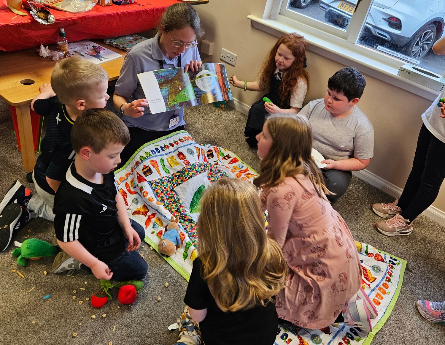 An adult dressed in a blue polo shirt holding a colourful picture book open in front of them. Seven children are gathered around a colourful cloth playmat in front of them looking at the pages, sitting on the floor in a semi circle surrounded by scattered toys.