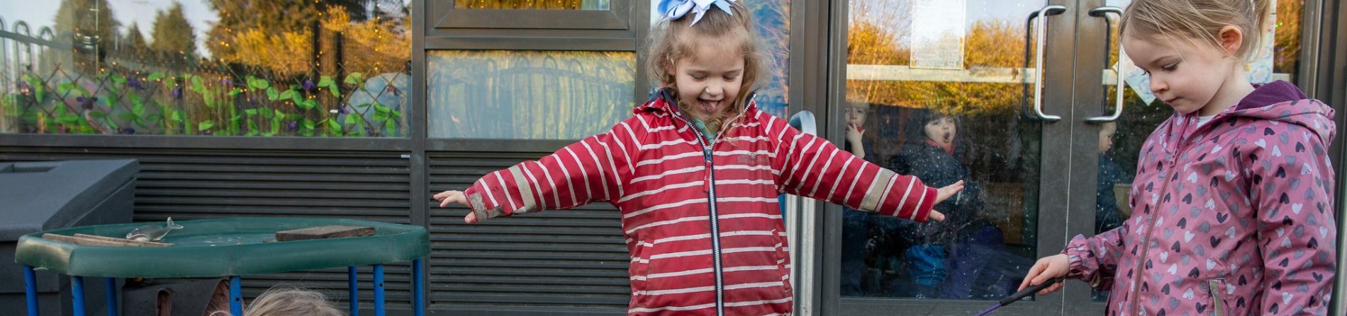 A child wearing a red striped coat is outside the Indigo Childcare Nursery, joyfully jumping and smiling