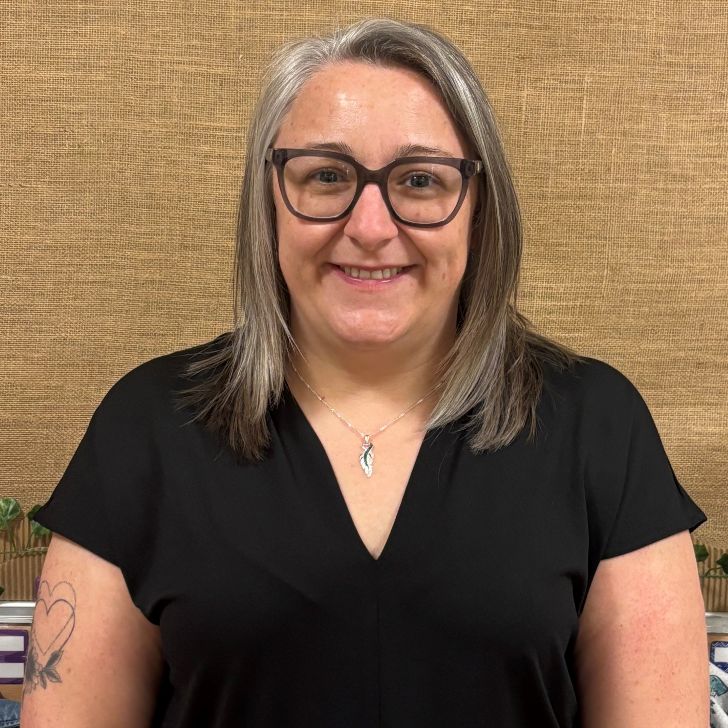Michelle smiles warmly for the camera. They are a white person with shoulder-length grey hair, wearing a black blouse and black glasses.