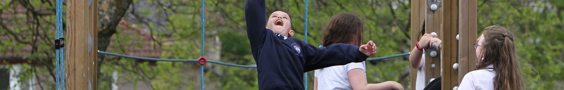 Four children dressed in school uniforms, playing happily outdoors on a wooden and rope climbing frame.