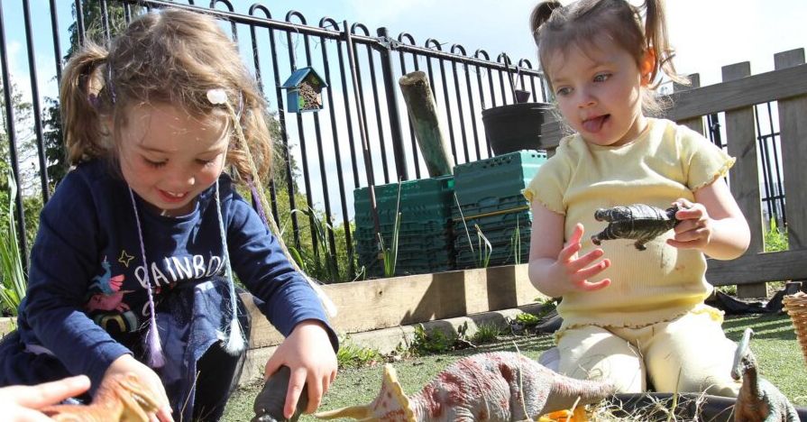 Two children kneeling on the ground outdoors, smiling and playing with dinosaur toys with a fence and blue sky in the background.
